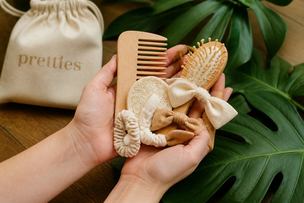 Close-up of hands holding various biodegradable hair accessories made from bamboo, wood, and organic cotton, arranged on a wooden table with green leaves in the background