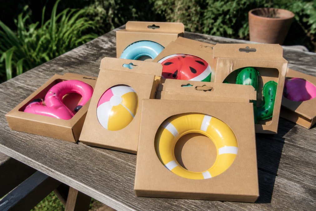Close-up of colourful inflatable pool toys still in their original cardboard and plastic-free packaging arranged on a weathered wooden garden table, warm afternoon sunlight casting soft shadows