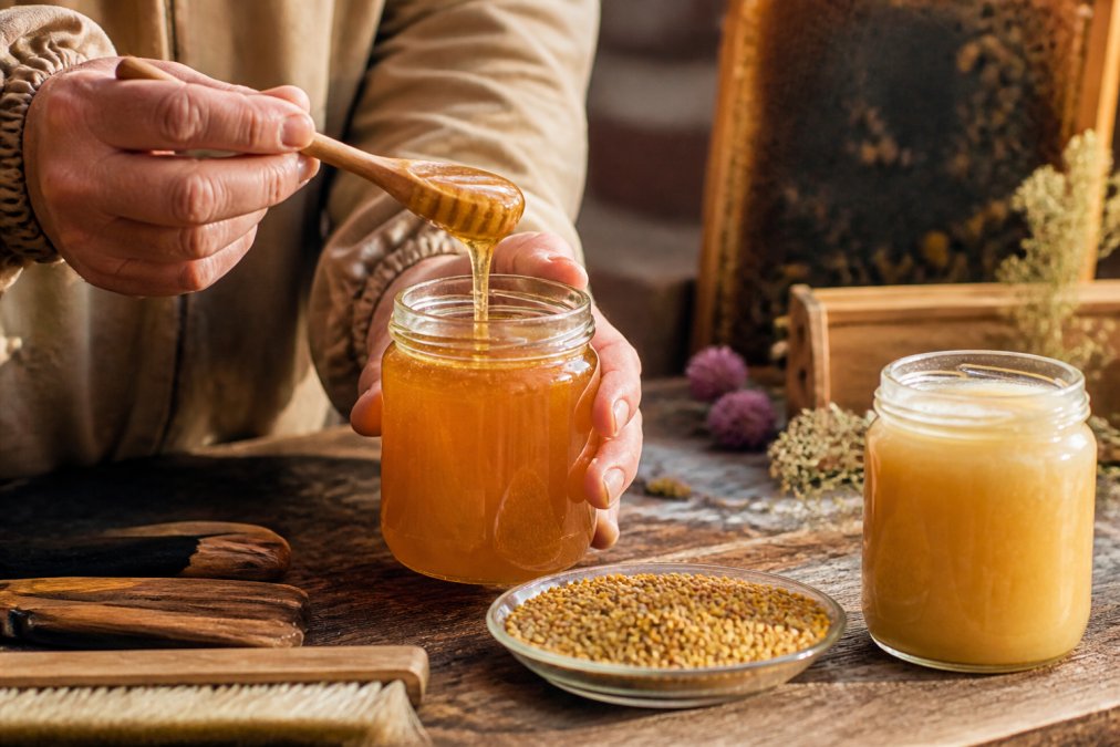 Nahaufnahme von Händen, die ein Glas goldgelben Honig mit Holzlöffel über einem rustikalen Holztisch halten, daneben Blütenpollen und ein Glas cremiger Sommerblütenhonig, warmes Tageslicht von der Seite, ländliche Imkerei-Atmosphäre