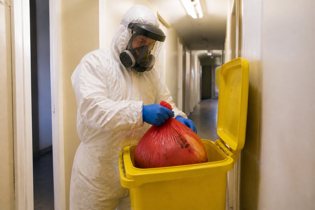 A fully suited biohazard technician in a white Tyvek coverall, nitrile gloves, respirator mask and face shield, carefully placing sealed red biohazard waste bags into a yellow clinical waste container inside a residential hallway under fluorescent lighting
