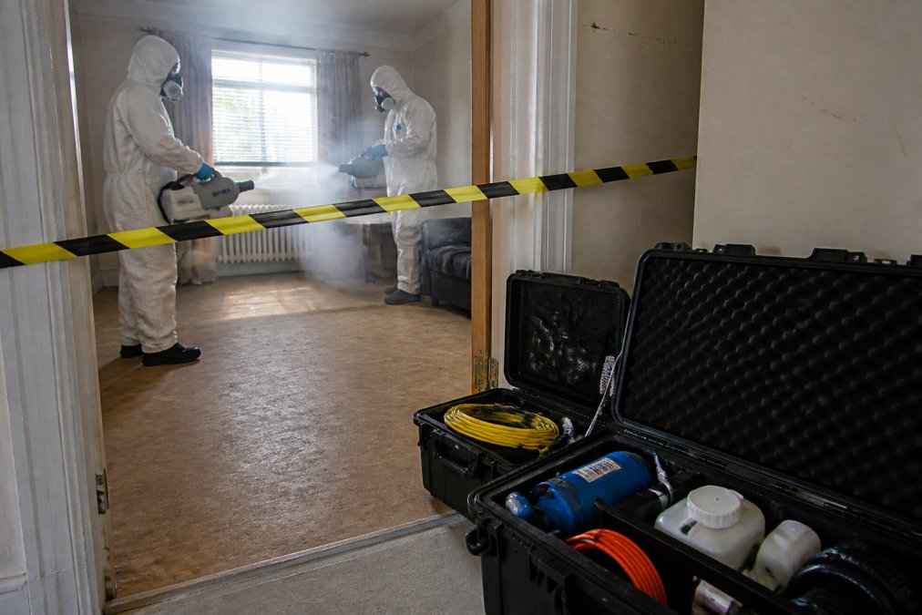 Two specialist cleaning technicians in full white Tyvek PPE suits using professional fogging equipment to decontaminate a room, with yellow hazard tape across the doorway and specialist cleaning equipment cases open in the foreground under bright work lighting