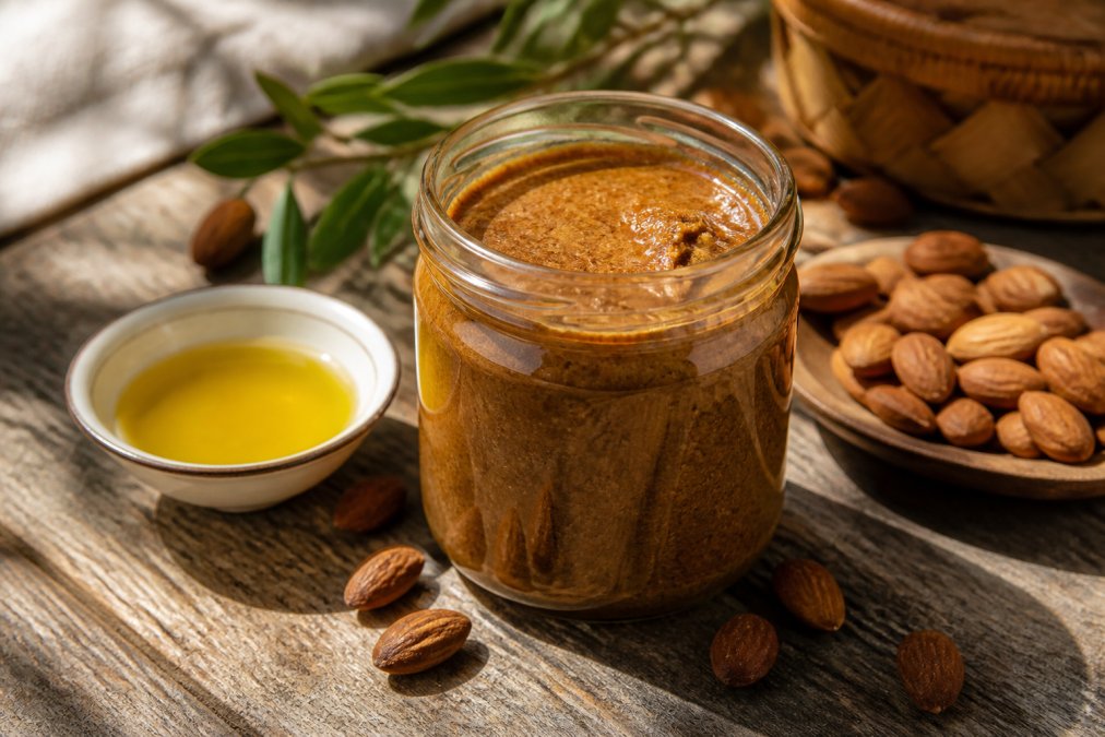 Een open glazen pot ambachtelijke amlou op een houten tafel, met geroosterde amandelen en een klein schaaltje gouden arganolie ernaast, warm natuurlijk daglicht