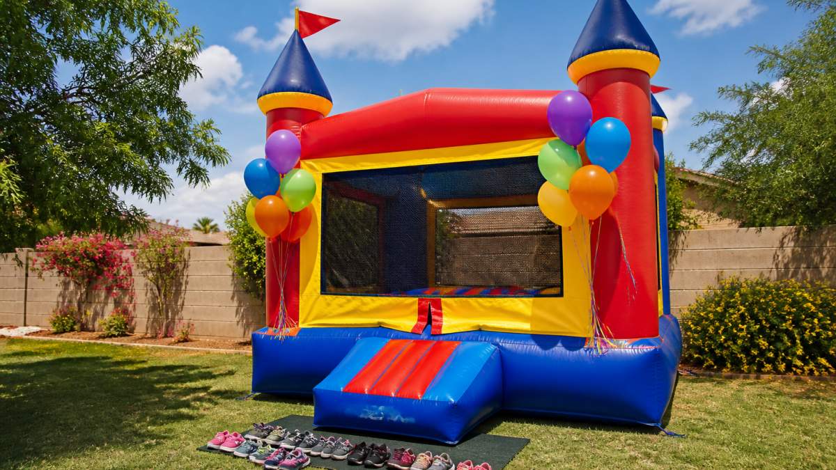 A colorful bounce house set up in a sunny backyard with balloons tied to the entrance and a birthday banner overhead, children's shoes lined up neatly on the grass beside it