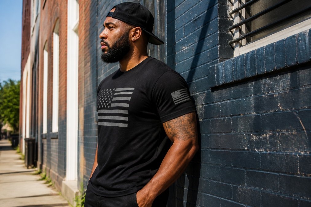 Person wearing a graphic t-shirt featuring a black american flag design while standing against a brick wall.