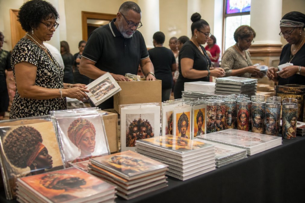 Diverse group of community members at a church gathering arranging and displaying stacks of Black heritage greeting cards, calendars, and cultural gift products on tables with warm interior lighting