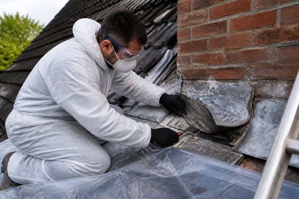 Professionele dakdekker in volledige veiligheidsbescherming, handschoenen, FFP2-masker, veiligheidsbril en beschermende overall, verwijdert voorzichtig oud bladlood van dakconstructie met zachte gereedschappen, terwijl plastic beschermingsfolie onder het werkgebied ligt