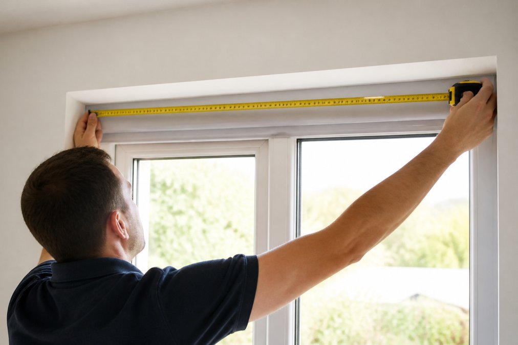 Homeowner using a metal measuring tape on a window frame, measuring horizontally at the top of the recess with the tape pulled taut, showing proper positioning and technique in natural daylight