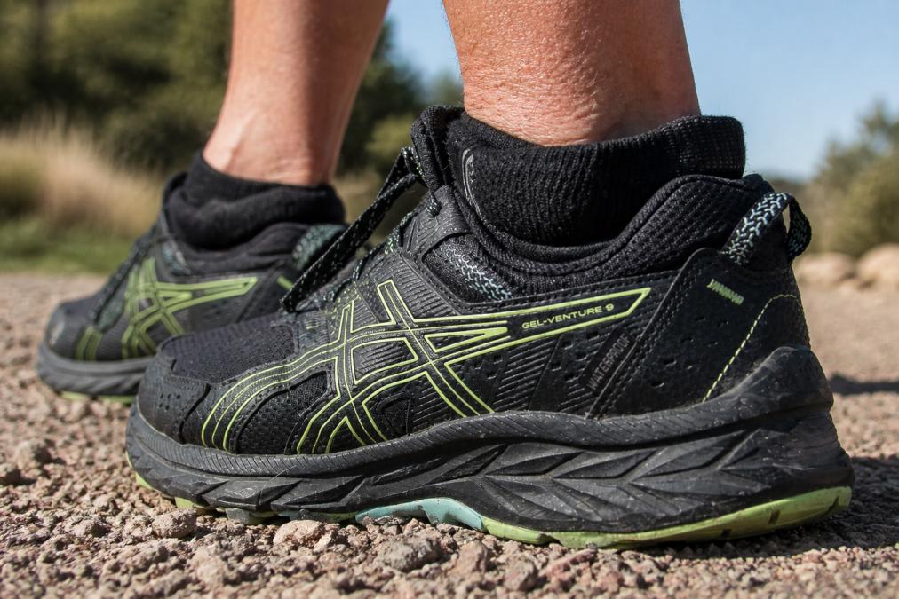 Close-up of a hiker's foot in a hiking boot on rocky trail terrain, showing proper sock fit and debris-resistant polyurethane barrier at the ankle collar preventing trail debris entry