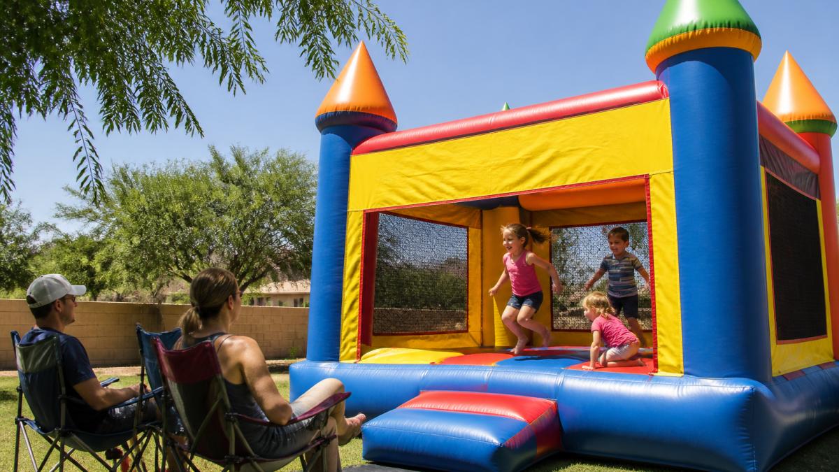 Children playing and laughing inside a colorful bounce house at an outdoor birthday party, with parents supervising from nearby lawn chairs on a bright sunny day