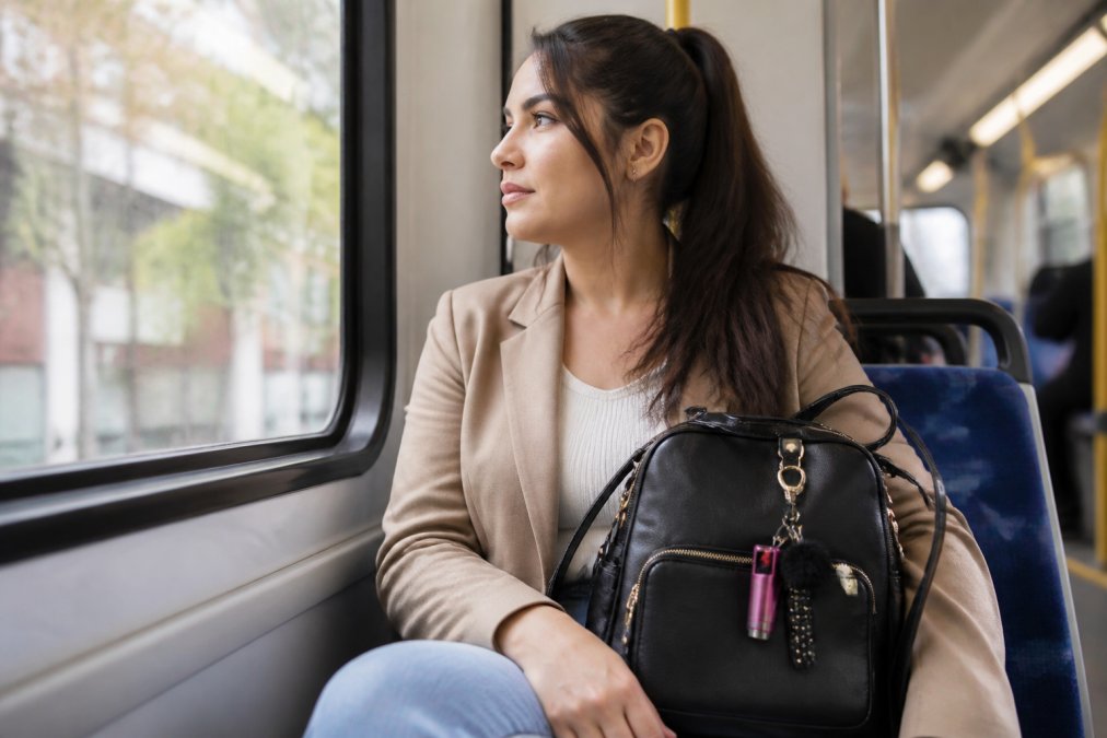 Young professional woman commuting on public transit with a stylish safety keychain attached to her bag's front pocket, natural daylight from transit window