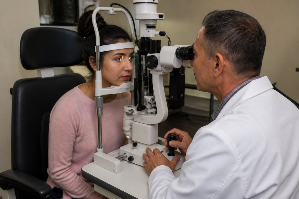 Patient sitting in an eye examination chair while an ophthalmologist performs a dilated eye exam using specialized equipment in a modern clinical setting