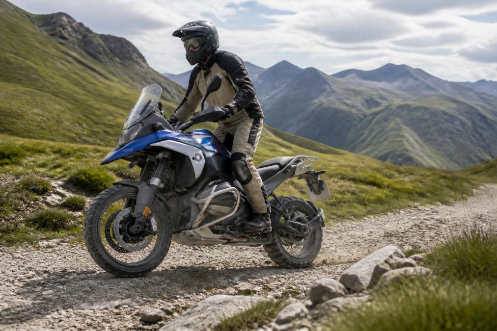 BMW R 1300 GS op een grindpad in berglandschap, zijkant zichtbaar met volle vering en banden in contact met onregelmatig terrein, demonstrerend het offroad-vermogen