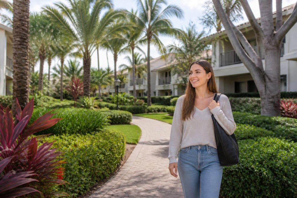 Young graduate student walking through lush, landscaped apartment community with mature palm trees and winding garden pathways, smiling and appearing relaxed on a sunny afternoon