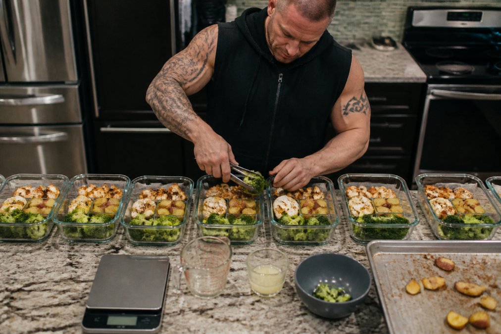 Seven meal prep containers with chicken, potatoes, and broccoli next to a digital food scale on a kitchen counter.