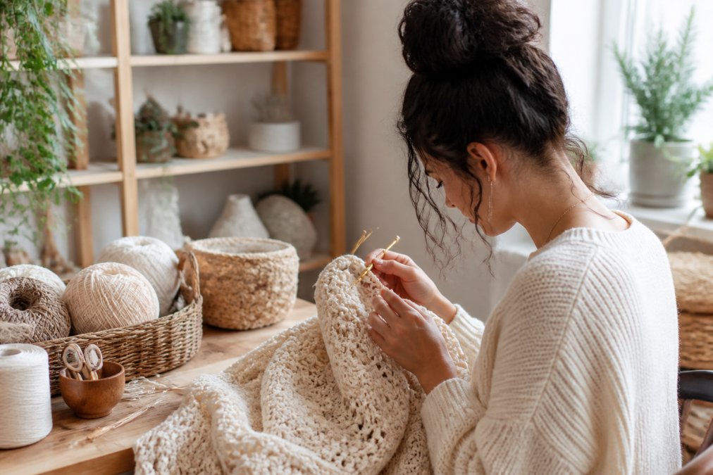 Close-up of a woman's hands crocheting a soft cream-colored baby blanket in a bright, cozy studio with yarn skeins and wooden tools nearby