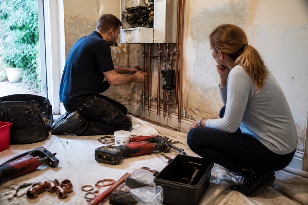 Professional installer working on boiler pipework and flue installation with tools and materials visible on the floor of a residential property, demonstrating the complexity of the work