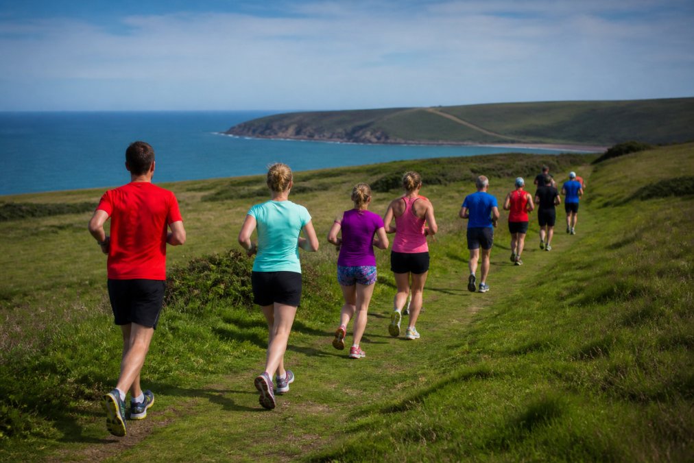 A group of runners in colourful kit jogging along a grassy coastal path on Bolberry Down, with the sea visible in the background under a bright morning sky