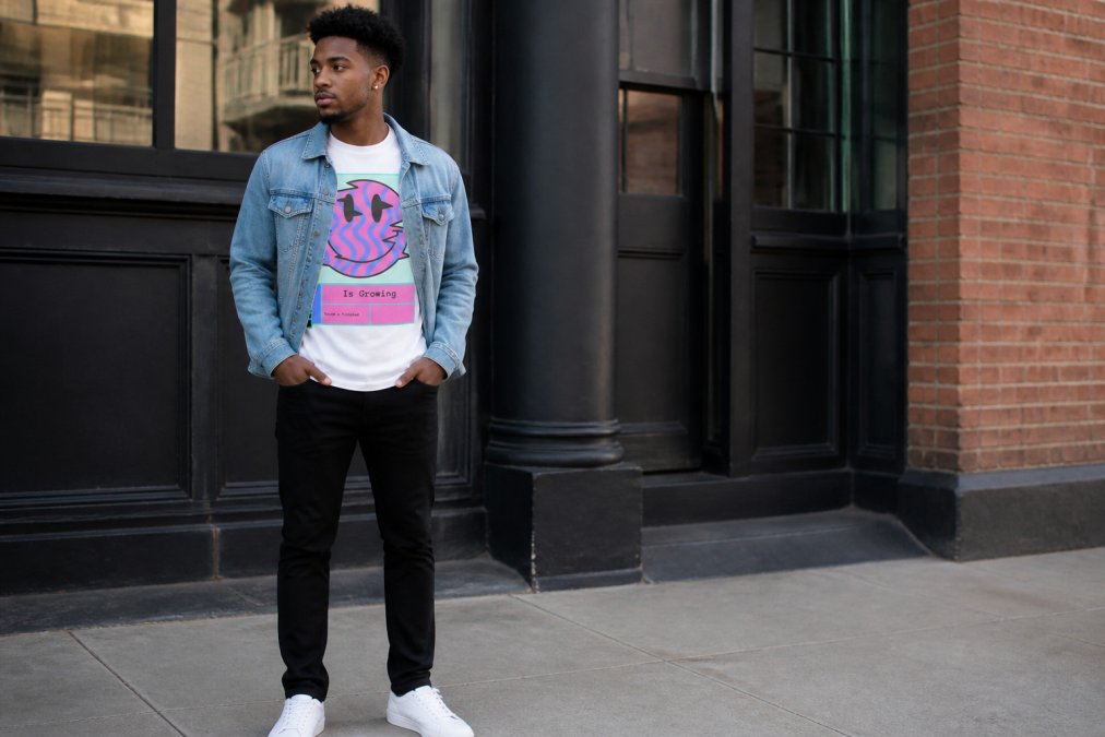 Young adult wearing a bold faith graphic tee layered under a structured denim jacket, styled with fitted black jeans and white sneakers, standing in modern urban setting with natural daylight