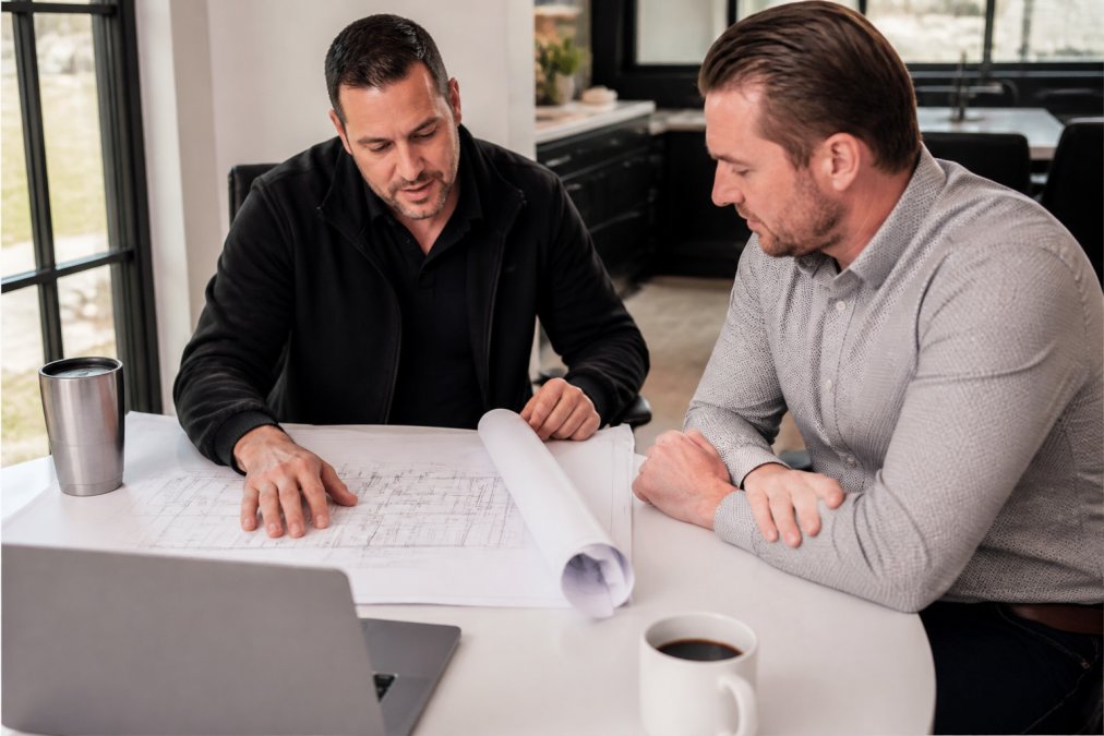 A general contractor and a commercial client reviewing blueprints and project plans together at a table in a bright office, with a laptop and coffee cups nearby