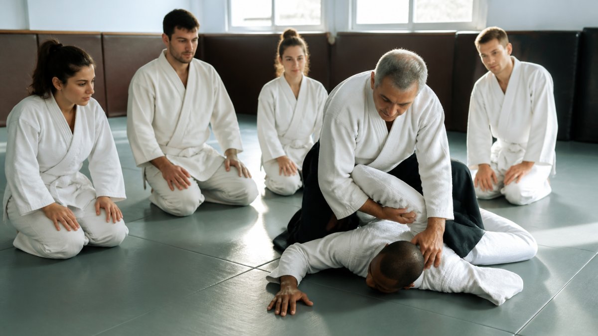Instructor demonstrating martial arts technique to a diverse group of beginner students in a bright, clean dojo with padded mats, natural lighting streaming through windows