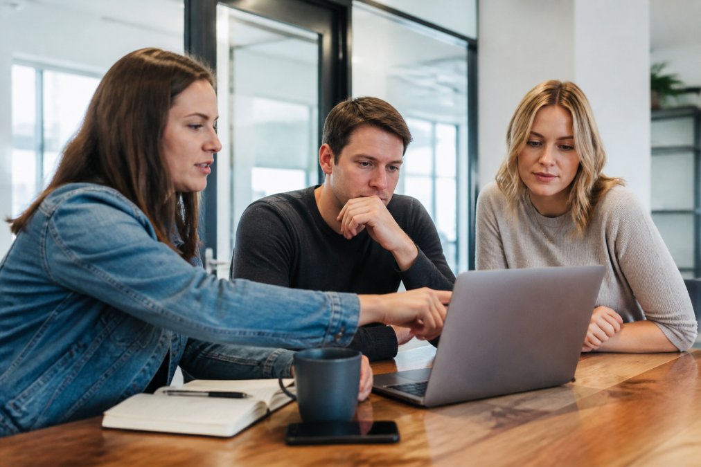 Small business team of three gathered around a laptop in a bright modern office, one person pointing at a screen showing a CRM pipeline dashboard, collaborative and focused atmosphere