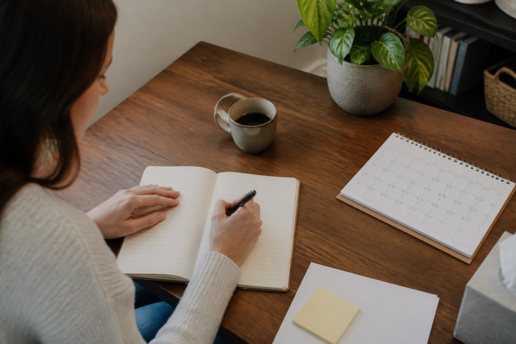 Person sitting at a desk with a notebook, writing down symptoms and medical history before an appointment, with a cup of coffee and calendar visible