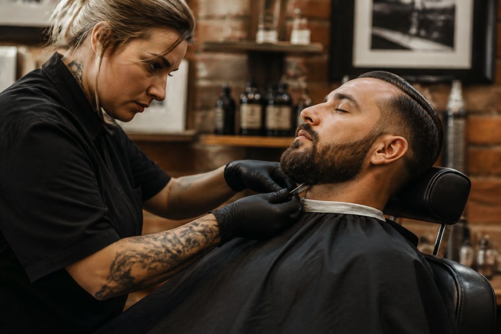 Professional barber carefully shaping client's beard with precision straight razor, focused expression, warm studio lighting highlighting beard texture and grooming detail