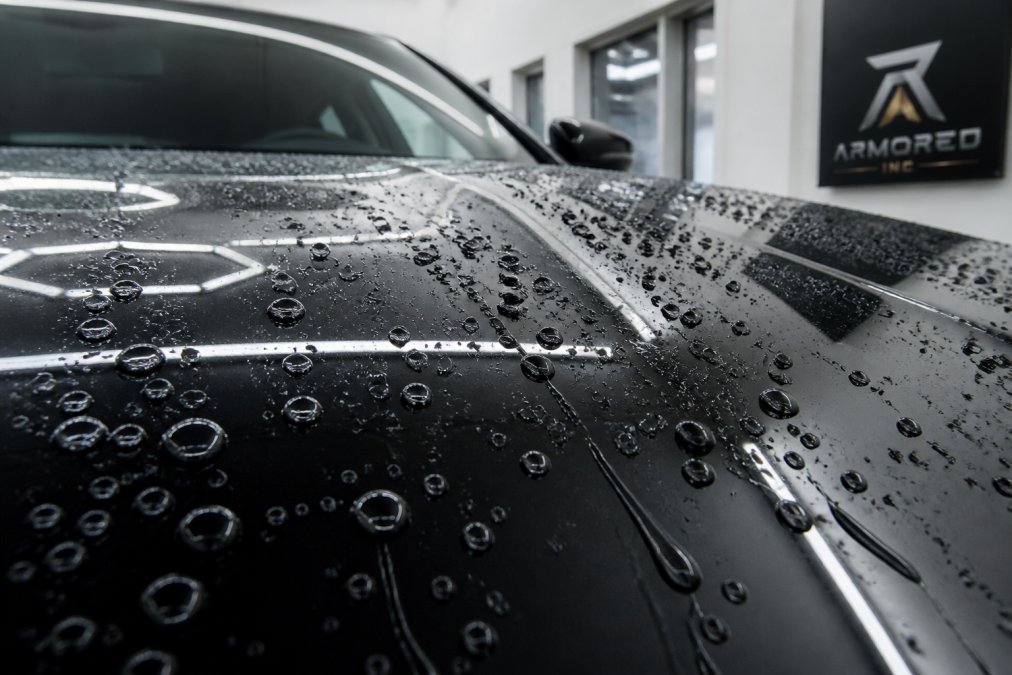 Close-up of a freshly ceramic-coated luxury vehicle's paint surface showing water droplets beading and rolling off the glossy finish, with dramatic light reflection highlighting the hydrophobic effect
