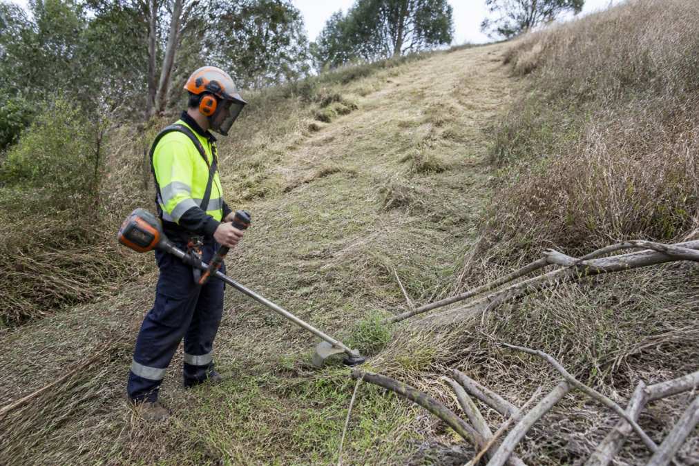 Worker performing commercial grounds maintenance by clearing dry grass to reduce fire hazards on a sloping property.