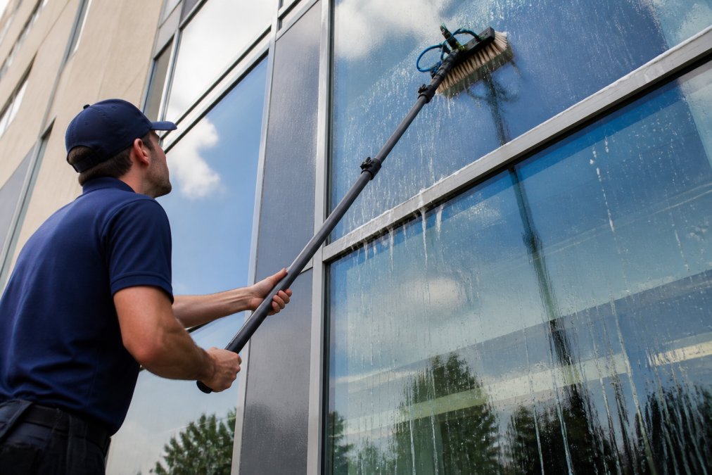 Close-up of a technician in a branded uniform operating a water-fed pole system with a brush head against a large commercial glass window panel, with soapy water running down the glass on a sunny day