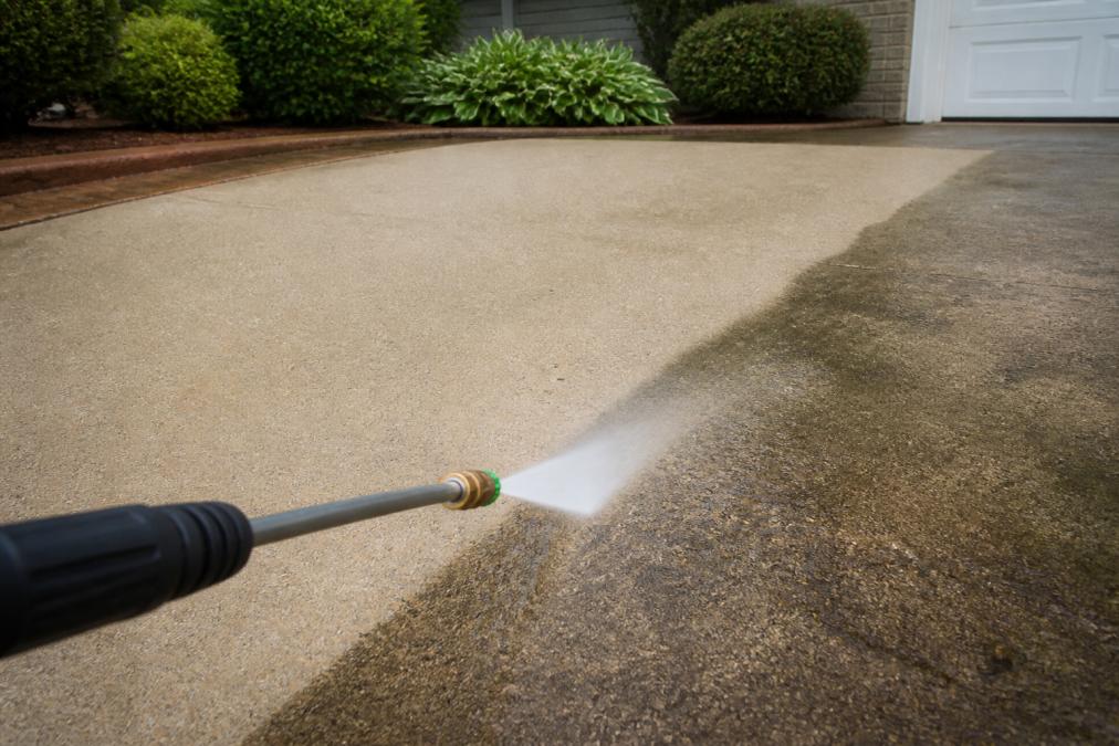 A clean concrete driveway with a visible contrast line between the freshly washed surface and a dirty section, with a professional-grade pressure washer wand in the foreground