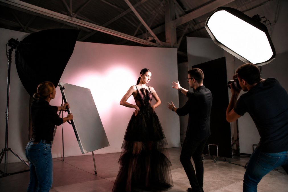 Fashion editorial shoot in progress with photographer directing model on set, professional lighting rigs overhead, assistants adjusting reflectors, creative energy visible on the studio floor