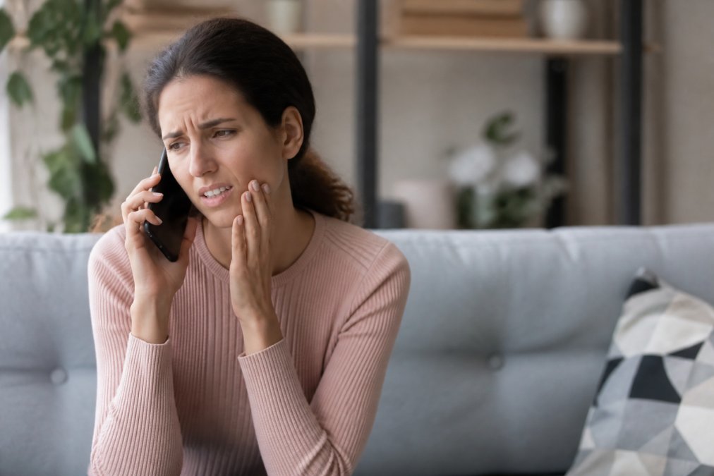 Patient sitting at home holding jaw in pain while making urgent phone call to dental office, concerned expression, natural indoor lighting