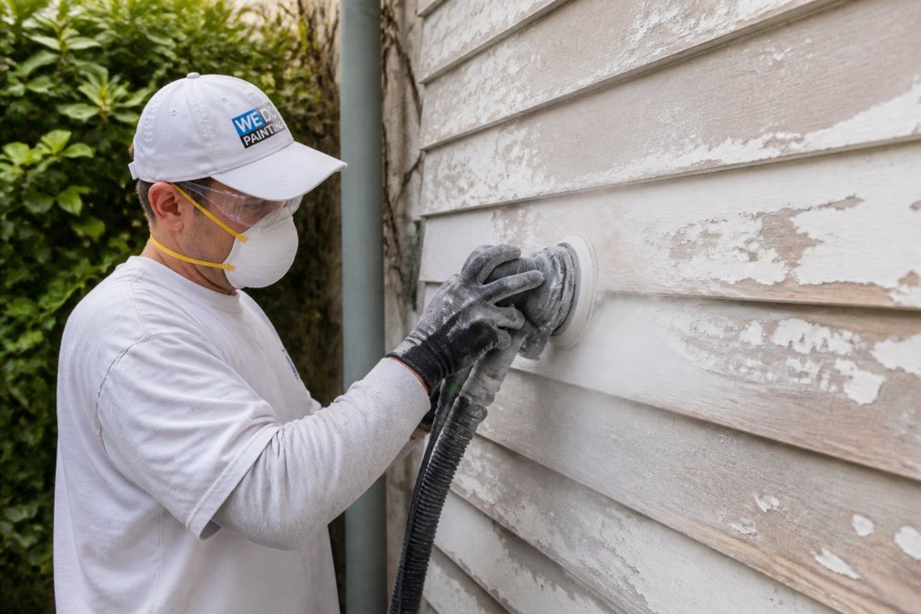 Professional painter in safety gear sanding weatherboard exterior wall with orbital sander, showing preparation dust and worn paint surface on residential home under natural daylight