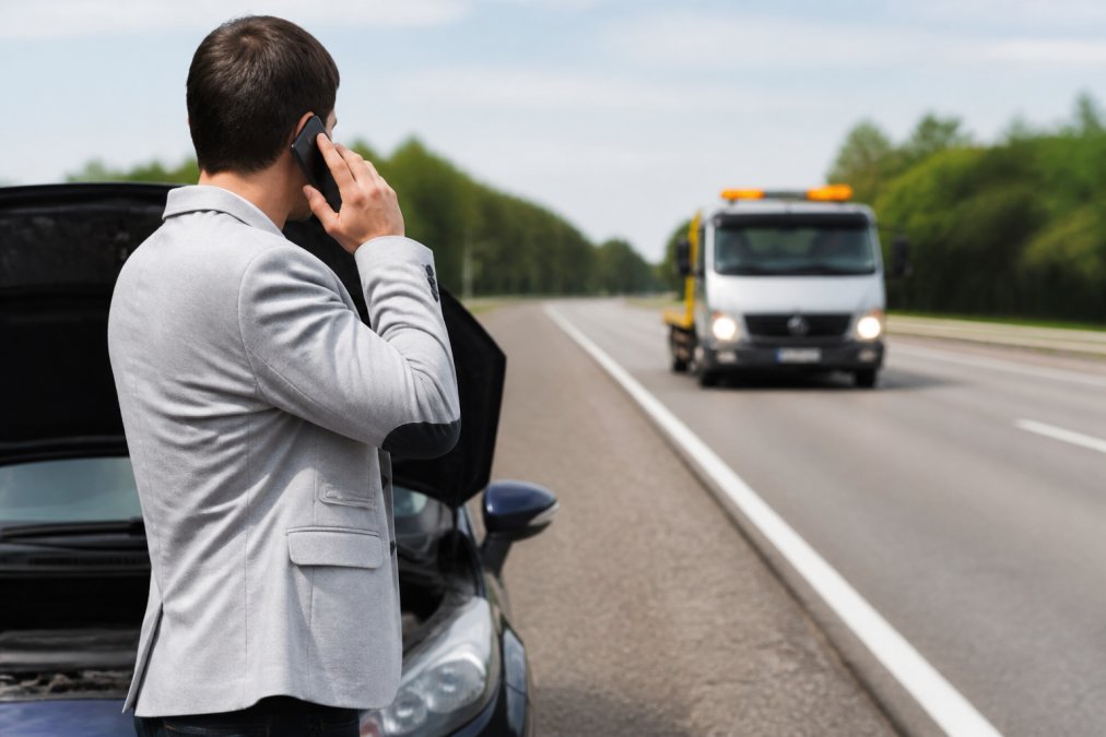 Driver standing safely beside their damaged vehicle on the roadside, using a mobile phone to call for emergency assistance, with a tow truck visible in the distance approaching on the highway