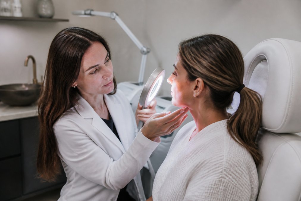 Board-certified dermatologist consulting with patient in modern medical spa treatment room, reviewing skin assessment with professional lighting and examining skin closely