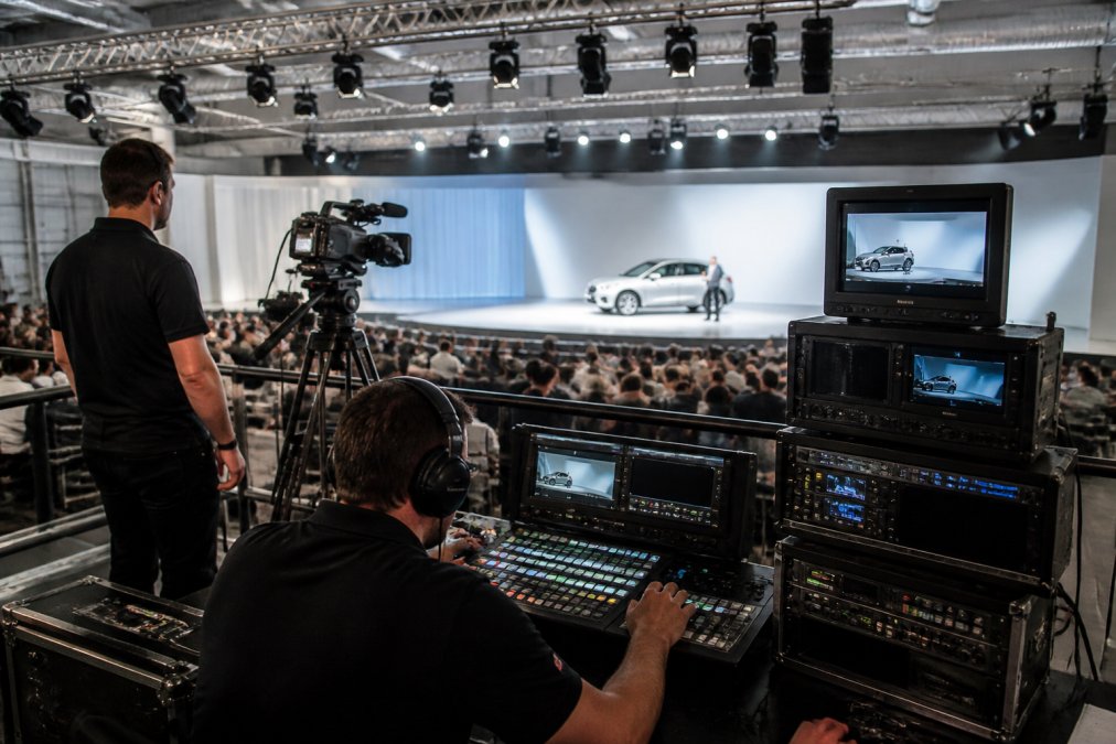 A professional live streaming crew setting up broadcast-grade cameras and monitors in a modern event venue, with a lighting rig overhead, a mixing desk in the foreground, and a producer reviewing feeds on a multi-screen rack