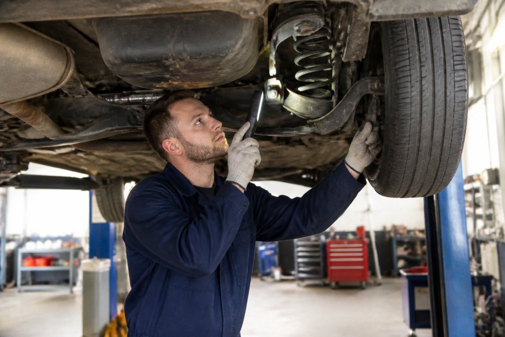 Professional mechanic in blue uniform performing a vehicle inspection under a car on a lift in a well-lit garage workshop, using diagnostic equipment and checking suspension components