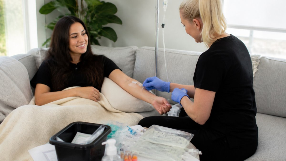 Professional registered nurse administering IV infusion to patient seated comfortably on home couch with medical equipment and supplies on nearby table, natural window lighting