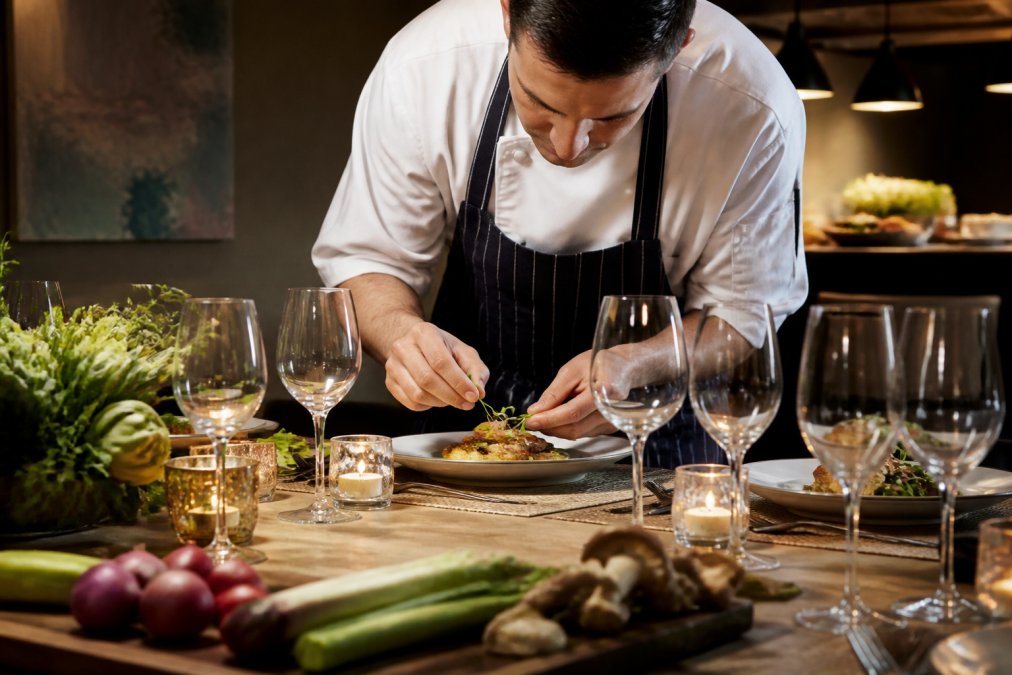 Private chef plating an elegant dish at a dinner table set with fine dining place settings, soft lighting, and fresh seasonal ingredients visible in the foreground