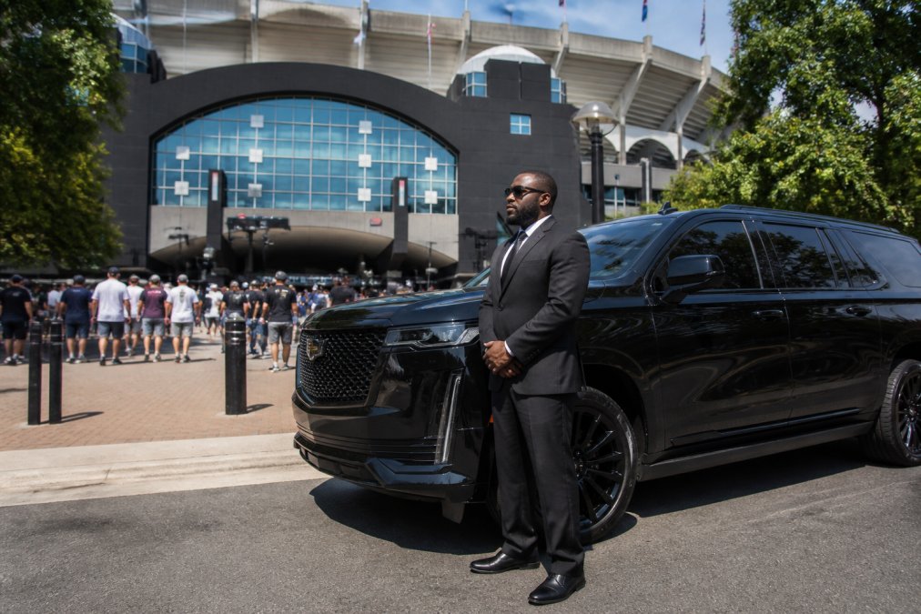 Professional driver in dark suit standing beside a luxury black SUV outside Bank of America Stadium, with game day crowds and stadium signage visible in the background