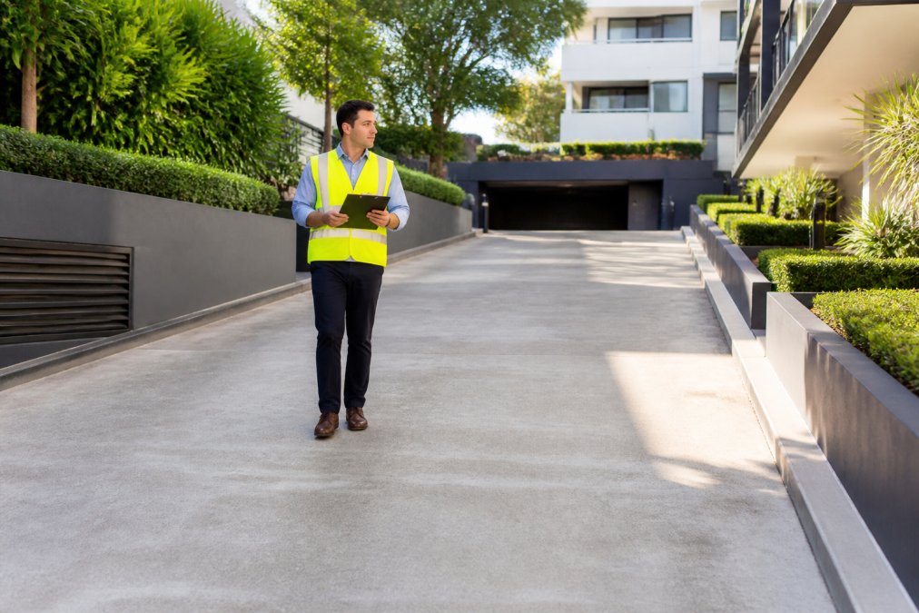 A facility manager in a high-vis vest walking a strata building's common driveway with a clipboard, checking a freshly cleaned concrete path and garden edging in bright morning light