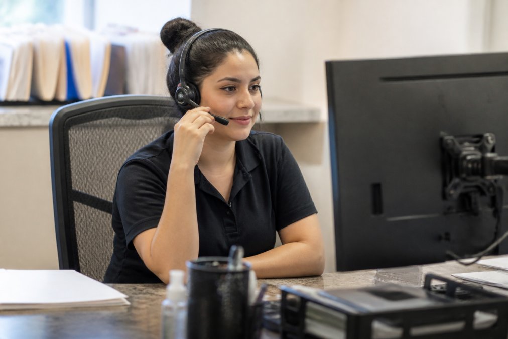 A friendly plumbing dispatcher at a desk with a headset, reviewing a service schedule on a computer screen in a bright office, taking an incoming call