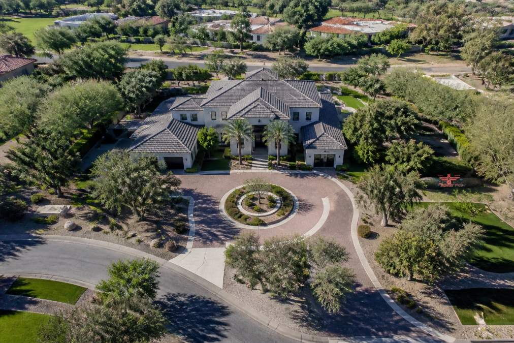 Aerial view of a high-end residential property showing the complete roofline, manicured landscaping, circular driveway, and surrounding neighborhood context from approximately 150 feet above ground