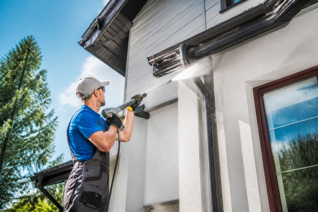 Professional technician in safety gear using pressure washing equipment on residential home exterior with water spray visible, bright daylight
