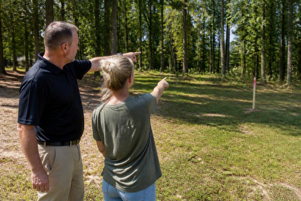 Homeowner and professional fence contractor walking the property perimeter together, both pointing at the yard where a fence will be installed, with trees and property boundary markers visible in the background