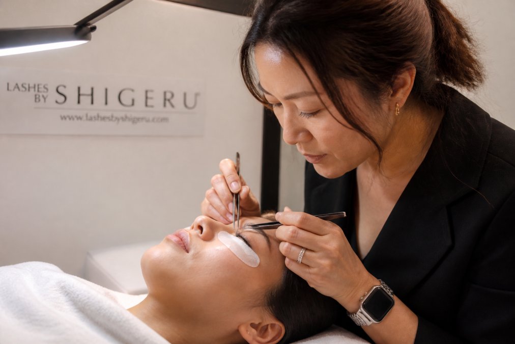 Professional lash technician carefully applying multiple lightweight extensions to a client's natural lash using precision tweezers under bright clinical lighting