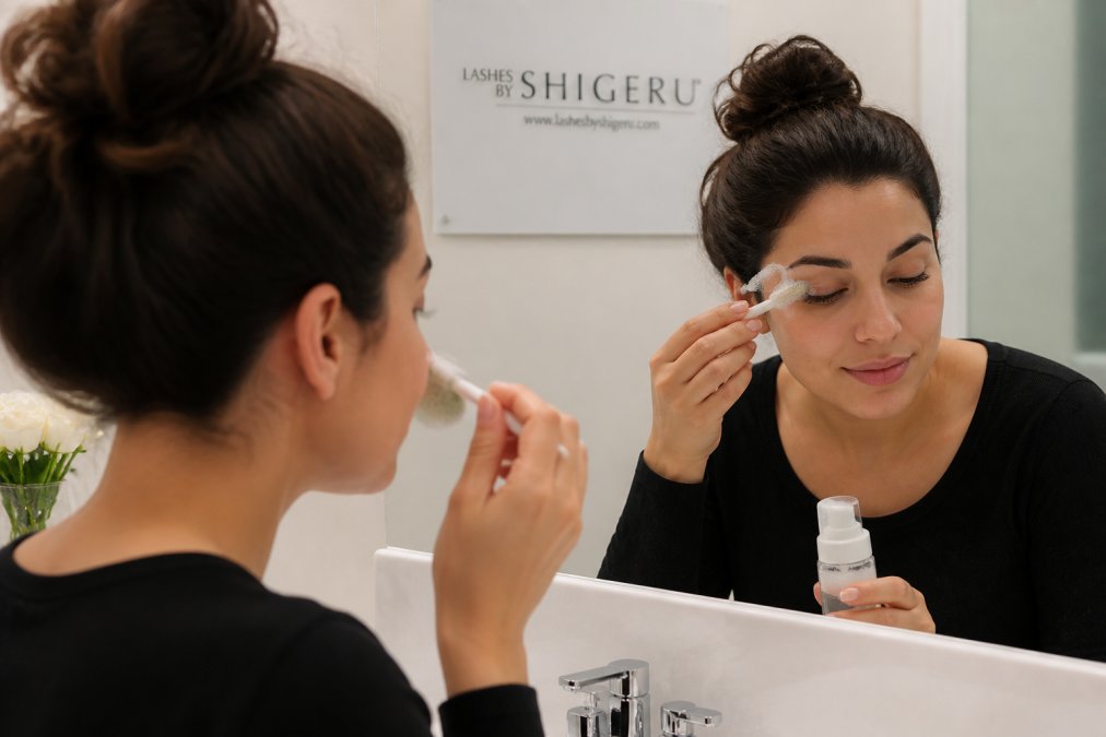Person gently cleansing eyelash extensions with an oil-free cleanser and soft brush, demonstrating proper daily maintenance technique over a bathroom sink with soft lighting