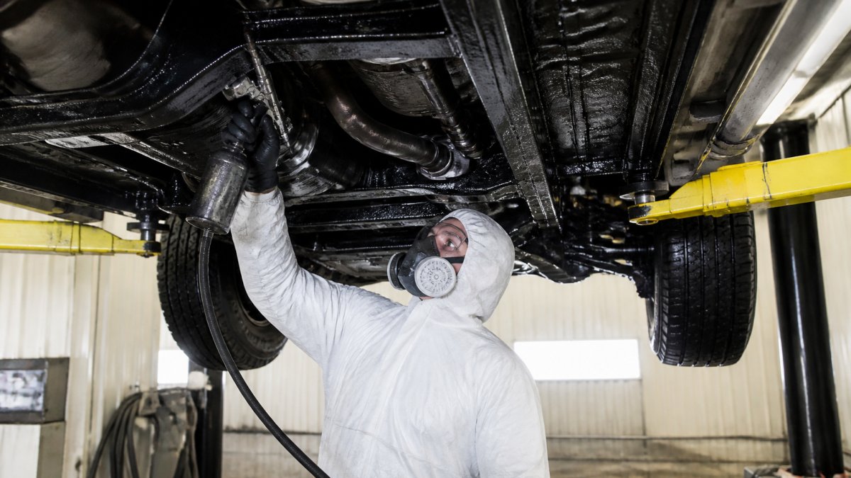 Professional technician in protective gear applying undercoating spray to vehicle undercarriage in shop bay, with spray equipment and metal frame visible under bright work lights
