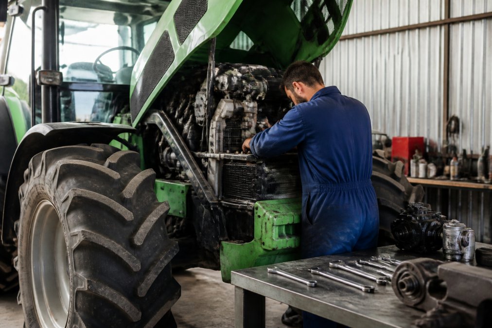 A diesel mechanic in blue overalls inspecting the engine bay of a large green 4WD tractor in a well-lit workshop, with spanners and spare parts arranged on a steel workbench in the background